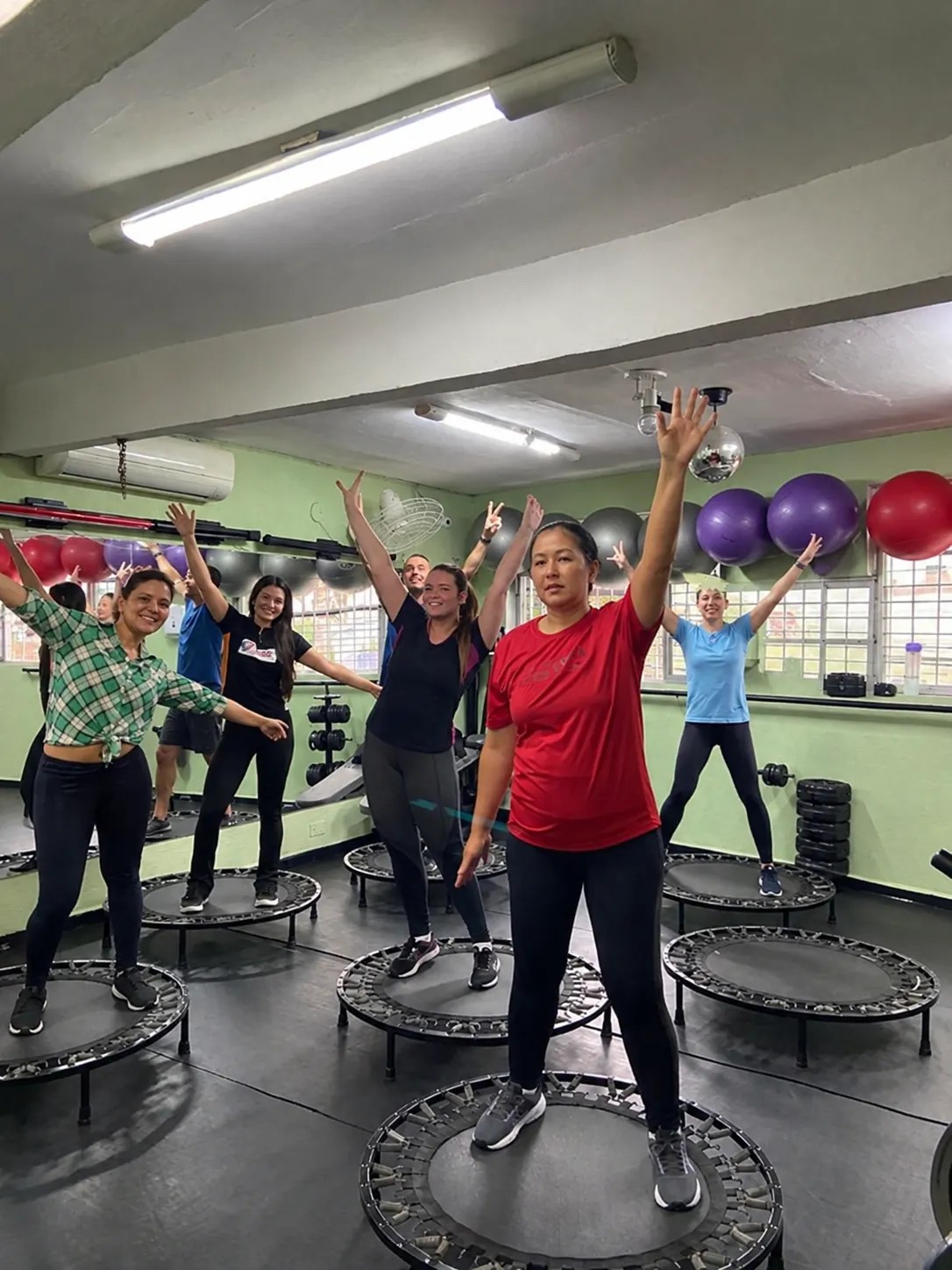 Alunas e alunos em pé nos minitrampolins, de braços abertos, na aula de jump
