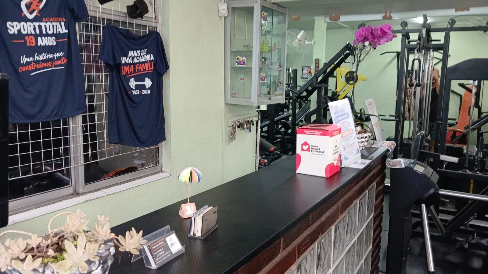 Recepção com balcão, camisetas dos 19 anos da academia e, ao fundo, os aparelhos de musculação